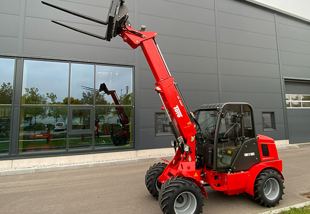 Toyo 850AT Teleskoplader mit Teleskoparm Roter Toyo 850AT Teleskoplader mit ausgefahrenem Teleskoparm und Palettengabel vor einer modernen Industriehalle mit Glasfront.