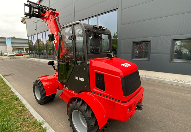 Roter Toyo 850AT Teleskoplader vor Firmengebäude Roter Toyo 850AT Teleskoplader mit Kabine und Palettengabel vor einem modernen Firmengebäude. Kompakter Lader für effiziente Arbeiten auf dem Betriebsgelände.