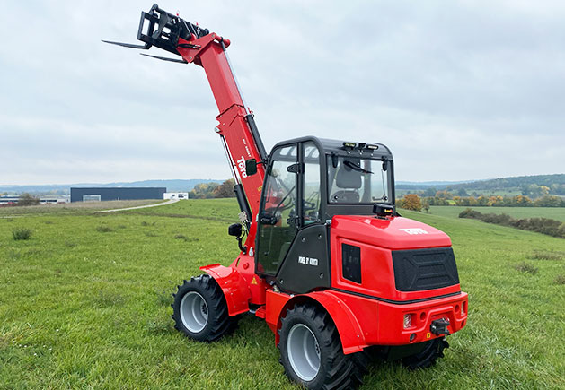 Toyo 850AT Teleskoplader mit Teleskoparm Roter Toyo 850AT Teleskoplader mit ausgefahrenem Teleskoparm und Palettengabel auf einer grünen Wiese vor einer ländlichen Kulisse.