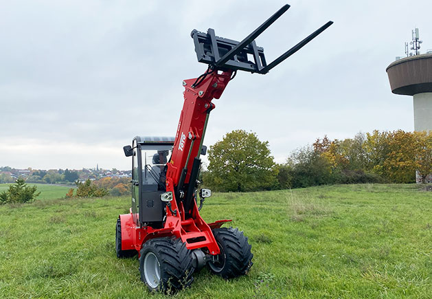 Toyo 850AT Teleskoplader mit Palettengabel Roter Toyo 850AT Teleskoplader mit ausgefahrenem Hubarm und Palettengabel auf einer grünen Wiese vor bewölktem Himmel und einem Wasserturm.