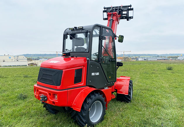 Roter Toyo 850AT Teleskoplader auf der Wiese Ein roter Toyo 850AT Teleskoplader mit angehobenem Ladearm steht auf einer grünen Wiese vor einer industriellen Kulisse.