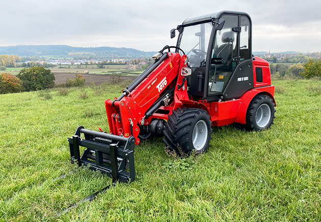 Toyo 850AT Teleskoplader im Einsatz Roter Toyo 850AT Teleskoplader mit Palettengabel auf einer grünen Wiese vor einer ländlichen Hügellandschaft bei bewölktem Himmel.