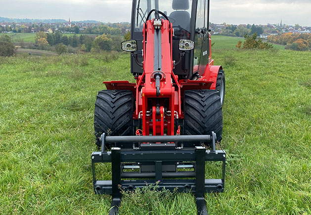 Roter Toyo 850AT Teleskoplader auf der Wiese Ein roter Toyo 850AT Teleskoplader mit Palettengabel steht auf einer grünen Wiese vor einer ländlichen Kulisse mit Bäumen und Häusern im Hintergrund.