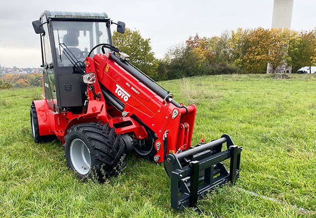 Toyo 850AT Teleskoplader im Einsatz Roter Toyo 850AT Teleskoplader mit Kabine und Anbaugerät auf einer grünen Wiese vor herbstlicher Kulisse.